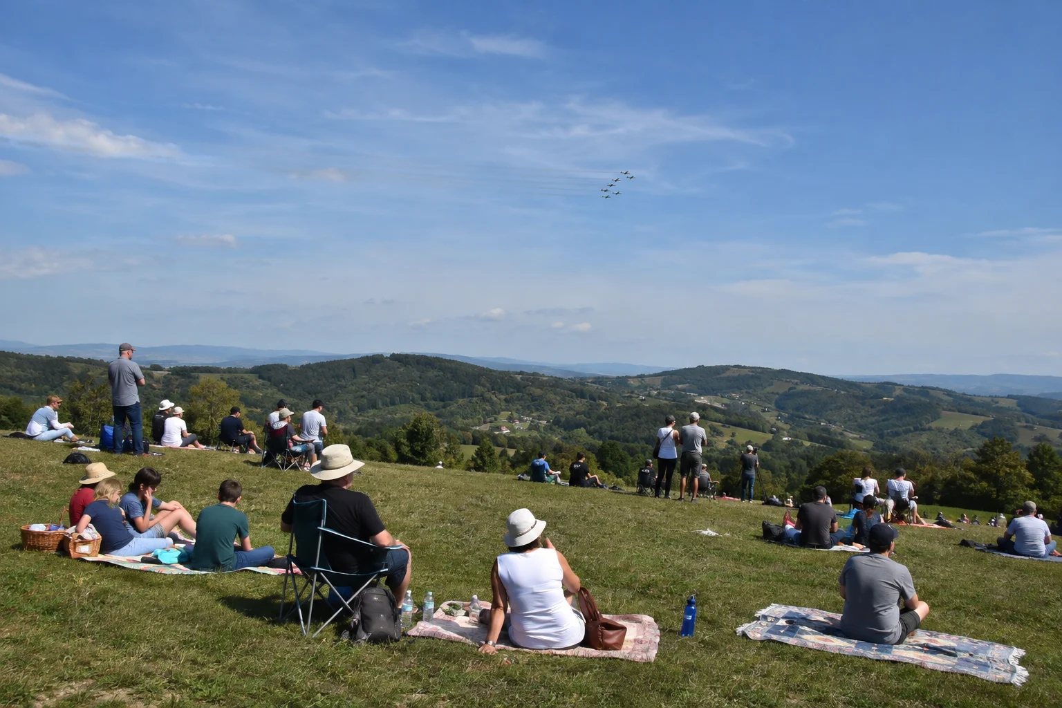 Besucher sitzen mit Decken und Wasser auf einer Wiese und beobachten Flugzeuge über dem Zagorje