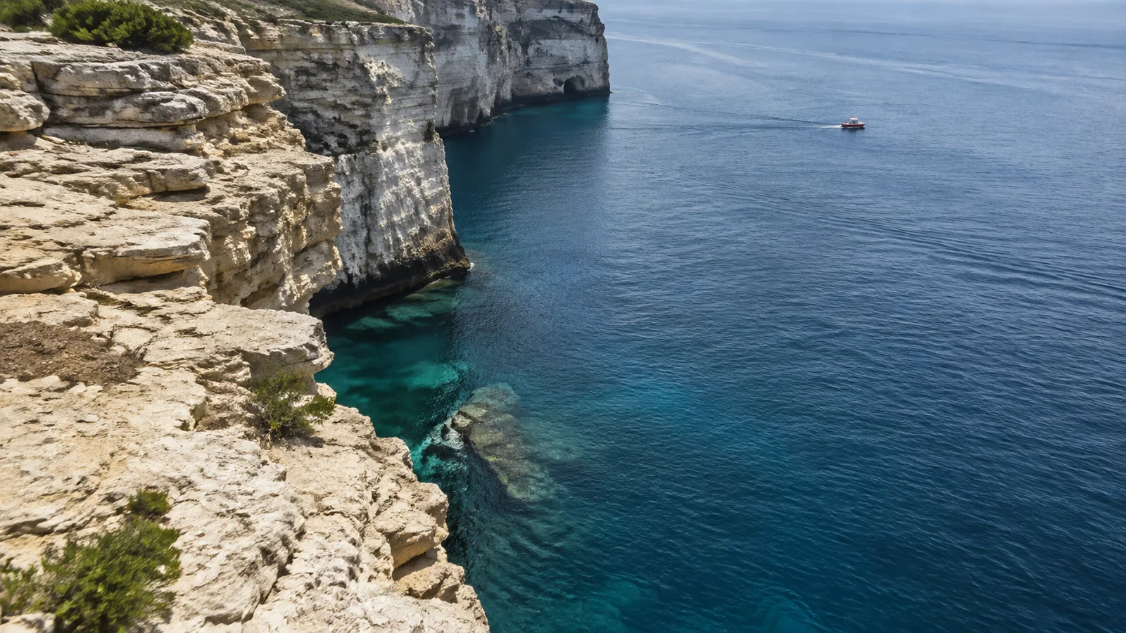 Hohe helle Felsen fallen bei Pula in tiefblaues Meer ab, ein kleines Rettungsboot liegt in der Ferne