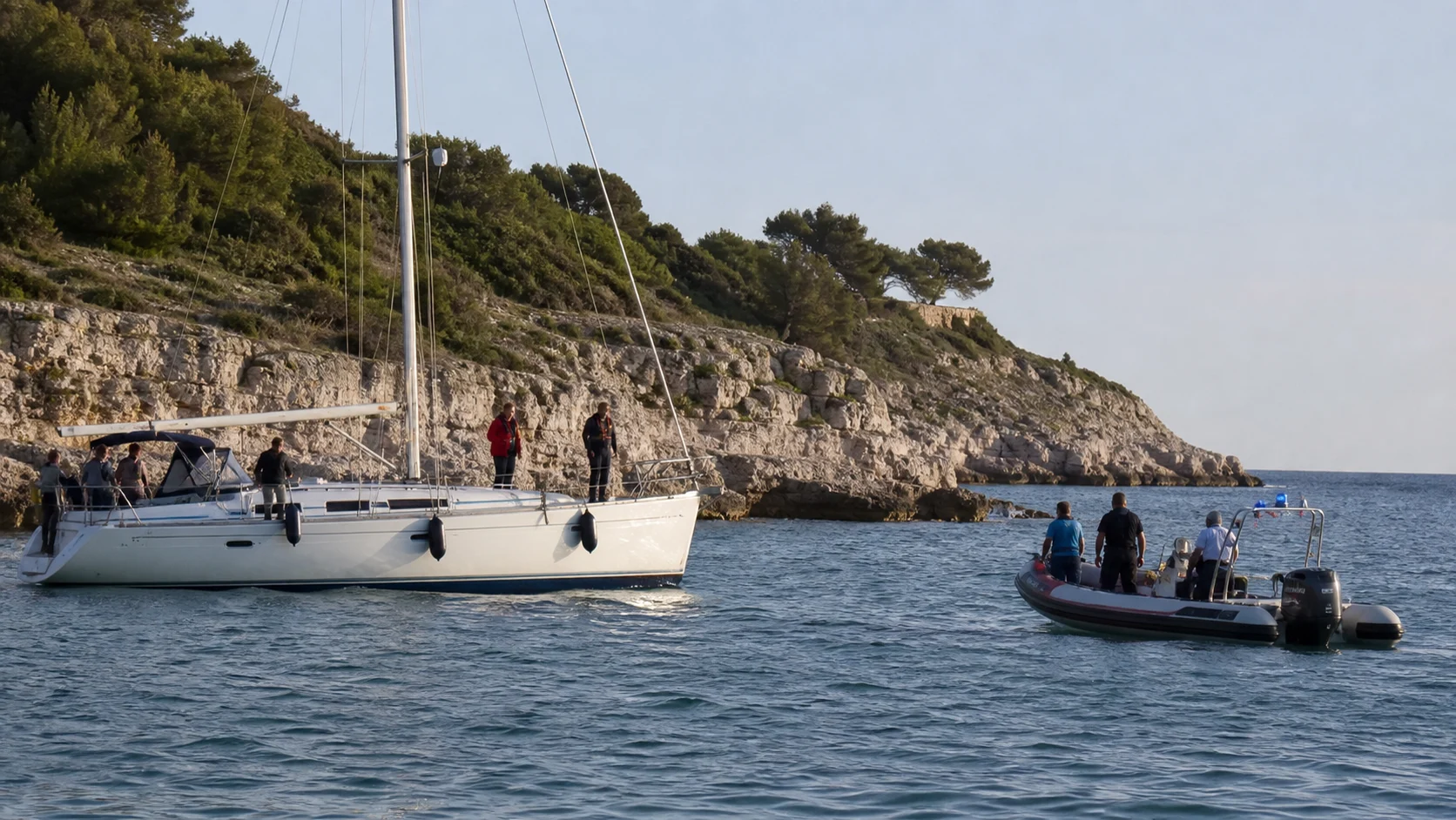 Eine gestoppte Segelyacht und ein Rettungsboot liegen vor einer felsigen Küste bei Pula