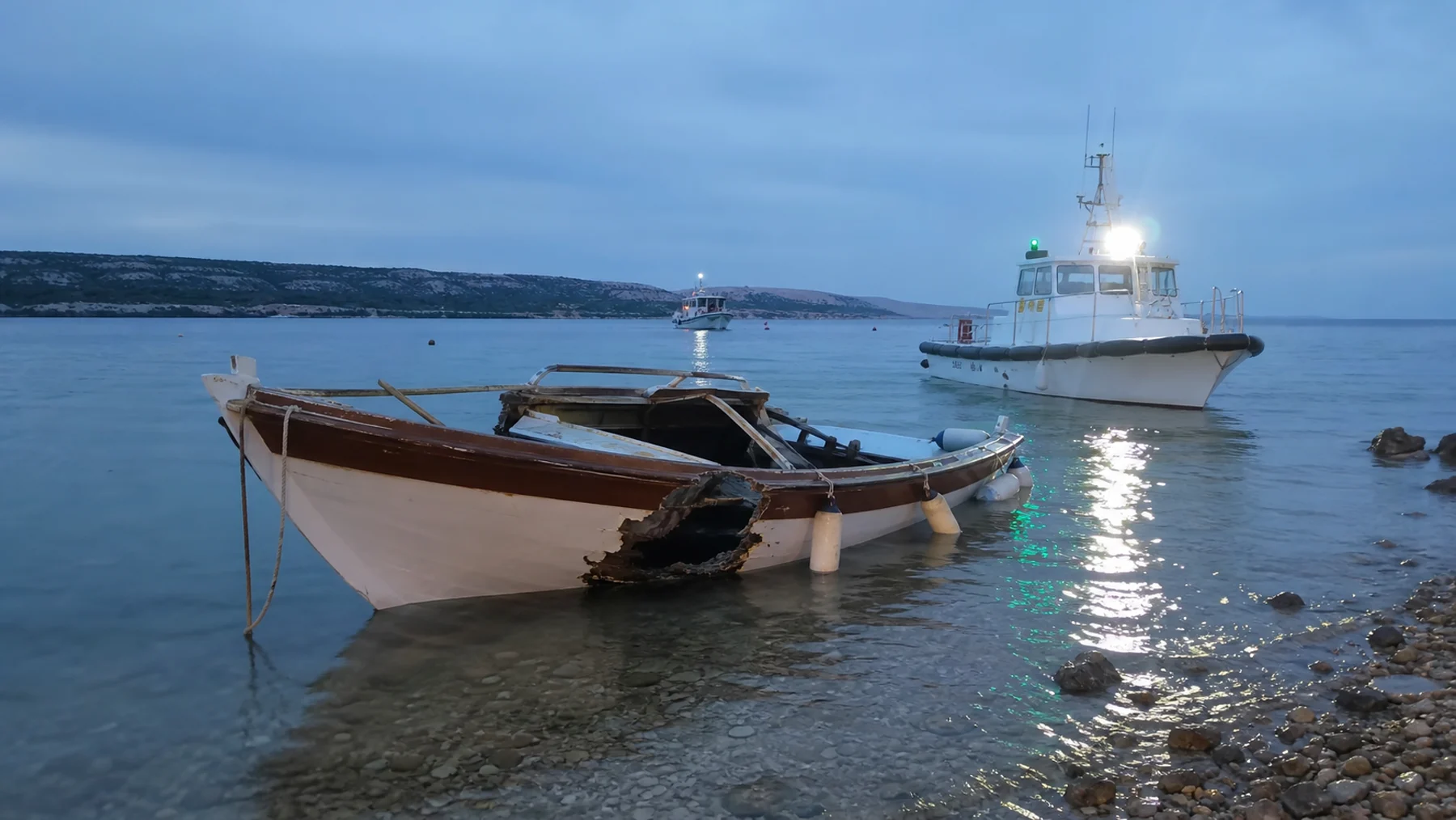Ein beschädigtes Holzboot liegt gesichert im Flachwasser, während ein Einsatzboot in der Nähe wartet